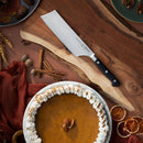 A pumpkin pie with whipped cream and pecans rests on a wooden tray next to the Messermeister Meridian Elite Nakiri Knife - 6.5", cinnamon sticks, dried orange slices, spices, and autumn decor—an essential fall kitchen scene.