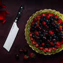 A vibrant fruit tart sits beside a Messermeister Meridian Elite Nakiri Knife - 6.5", surrounded by scattered cherries, a strawberry, and rose petals on a dark surface—a must-have tool from Messermeister.