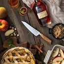 A table features a bottle of Maker’s Mark bourbon, a glass of whiskey, apples, apple pie, brown sugar, cinnamon sticks, sliced apples in a dish, and the Messermeister Oliva Elite O.T. Prep Knife - 6.5" ready for perfect slicing.