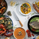 A top view of a table with lobster, oysters, shrimp, fish, gnocchi, and soup garnished with herbs and lemon wedges. At the center is the Messermeister Oliva Elite O.T. Prep Knife - 6.5", ready for expert preparation.
