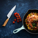 A skillet of shrimp and diced vegetables rests on a blue surface beside a Messermeister Oliva Elite O.T. Prep Knife - 6.5" with a wooden handle and a bunch of vine tomatoes.