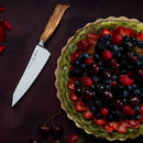 A fruit tart with strawberries, blueberries, cherries, and blackberries rests on a green plate. Next to it, a Messermeister Oliva Elite O.T. Prep Knife - 6.5" with a wooden handle lies on the dark red surface.