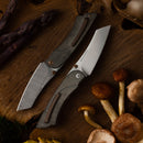 Two Messermeister Ojai Folder - 3.75" knives with textured handles and patterned blades rest on a wooden surface, surrounded by mushrooms and purple asparagus.