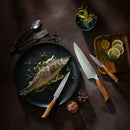 A cooked whole fish rests on a bed of greens atop a black plate, surrounded by lemon slices, herbs, seasonings, and three precision-forged Messermeister knives—all part of the Messermeister Oliva Elite 11-Piece Next Level Knife Block Set—displayed with pride near its sleek magnetic knife block on a dark surface, exemplifying the exceptional craftsmanship of Messermeister.