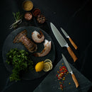 A flat lay showcases raw lobster tail, fresh herbs, lemon slices, and three knives from the Messermeister Oliva Elite 11-Piece Next Level Knife Block Set arranged on a dark surface. Surrounding the ingredients are bowls filled with spices, salt, pepper, and vibrant chopped peppers. The sophisticated kitchen setup is completed with a sleek Messermeister magnetic knife block.