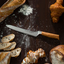 The Messermeister Oliva Elite Offset Scalloped Bread Knife - 8" with a wooden handle rests on a floured surface, surrounded by sliced and whole baguette, a croissant, a loaf of bread, and an iced pastry.