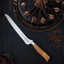 The Messermeister Oliva Elite Offset Scalloped Bread Knife - 8" with a wooden handle rests on a dark surface beside a glossy chocolate cake adorned with frosting swirls, chocolate curls, and scattered chocolate shavings.