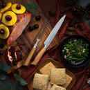 A glazed ham with pineapple and cherries is served on a wooden board beside green beans with almonds, square biscuits, pinecones, greenery, and the Messermeister Oliva Elite Kullens Carving Set - 2 Piece by Messermeister on a dark cloth.