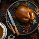 A roasted whole turkey is served on a round slate platter, garnished with herbs, dried orange slices, and cranberries, with a Messermeister Oliva Elite Kullens Carving Knife - 8" nearby. Pie, pinecones, and autumn leaves complete the scene.