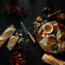 A charcuterie board sits on a dark table with cheese, figs, berries, meats, pickles, and honey, surrounded by baguettes, grapes, strawberries, and a jar of honey. Olive wood handle knives from the Messermeister Oliva Elite Magnet Block Set (6 Piece) are displayed nearby.