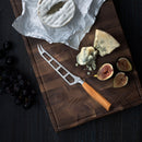 The Messermeister Oliva Elite Magnet Block Set – 6 Piece, featuring elegant olive wood handles, accompanies assorted cheeses, figs, and grapes on a wooden cutting board. One cheese wheel is partially unwrapped with crumbs scattered nearby.