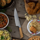 The Messermeister Oliva Elite 11-Piece Next Level Knife Block Set from Messermeister sits elegantly on a wooden table, surrounded by fried chicken, chips, a burger, baked beans, potato salad, and pie—a tableau celebrating both exceptional culinary craftsmanship and the comfort of hearty home cooking.