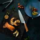 A cooked lobster tail rests on a cutting board with a chef’s knife—specifically a Messermeister Oliva Elite—surrounded by lemon wedges, fresh herbs, and roasted garlic. On the dark surface nearby are tongs, a blue plate, a fork, and tomatoes. The elegant Messermeister Oliva Elite 11-Piece Next Level Knife Block Set is showcased in the background on its magnetic knife block.