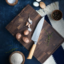 A wooden cutting board with a Messermeister Oliva Elite 11-Piece Next Level Knife Block Set chef’s knife, garlic cloves, potatoes, and fresh herbs. Surrounding the board are bowls of salt, pepper, flour, and eggs—all arranged on a dark blue surface with a woven cloth.