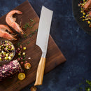 The Messermeister Oliva Elite Nakiri Knife - 6.5" with a wooden handle sits on a cutting board by raw shrimp, chopped herbs, tomatoes, and corn salad—perfect for vegetable prep on the dark blue surface.