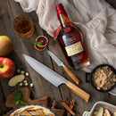 On a wooden table sits a bottle of bourbon, two knives from the Messermeister Oliva Elite 11-Piece Next Level Knife Block Set by Messermeister, a glass of whiskey, apples, sliced fruit, a bowl of brown sugar, cinnamon sticks, a pie, and a light cloth.