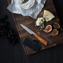 An elegant arrangement featuring a wooden board with a cheese knife, assorted cheeses (some crumbled), figs, grapes, and a black Messermeister knife case, with the knife resting at the center—an impressive display that would perfectly complement the Messermeister Oliva Elite 11-Piece Next Level Knife Block Set.
