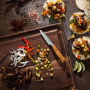A wooden board with chopped vegetables, sliced beef, a Messermeister Oliva Elite knife, and taco toppings sits beside assembled tacos, spices, and lime wedges on a dark table—perfectly complemented by the sleek Messermeister Oliva Elite 11-Piece Next Level Knife Block Set.