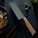 A Messermeister Oliva Elite 11-Piece Next Level Knife Block Set knife with a wooden handle rests on a dark textured table next to a bowl of noodles and vegetables, chopsticks, and a small dish of sliced green onions.