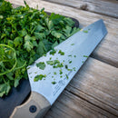 A Messermeister Overland Chef 6-Piece Leather Knife Roll Set chef’s knife, adorned with chopped parsley on a wooden cutting board, rests atop a rustic wooden table; scattered parsley pieces cling to the blade, highlighting the effortless precision of Messermeister’s quality outdoor cooking knives—freshly selected from the renowned Overland Chef Leather Knife Roll.