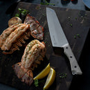 Grilled lobster tails with herbs and lemon wedges are served alongside the Messermeister Overland Chef Camp Knife - 8", crafted from Nitro-B steel, on a dark wooden cutting board.