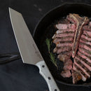 Sliced medium-rare steak with the bone rests in a black cast iron skillet, topped with sprigs of rosemary. A large chef’s knife, just taken from the Messermeister Overland Chef 6-Piece Leather Knife Roll Set, lies beside the skillet on a dark surface.