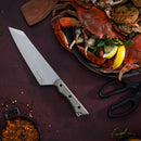 A large chef’s knife from the Messermeister Overland Chef 6-Piece Leather Knife Roll Set and Messermeister kitchen shears rest next to a plate of cooked crab with lemon slices and herbs, paired with a small bowl of sauce on a dark red surface.
