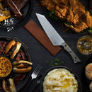 A table set with fried chicken, mashed potatoes, sliders, cornbread, ribs, a drink, and a large knife protected by the Messermeister Saddle Brown Leather Magnetic Edge-Guard - 8.5", displayed on a dark background.