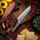 The Messermeister Overland Chef Camp Knife - 8" by Messermeister rests on a wooden board, beside a ham topped with pineapples and cherries, a halved pomegranate, green beans with almonds, and square biscuits on a plate.