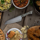 A Messermeister Overland Chef Camp Knife - 8" rests on a wooden table with bowls of baked beans, potato salad, fried chicken, chips, a hamburger, pie, herbs and salt. This versatile Messermeister camp knife features premium Nitro-B steel.