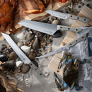 Four knives from the Messermeister Overland Chef 6-Piece Leather Knife Roll Set are arranged on sand among rocks, seashells, a cooked crab resting on a cutting board, a live blue crab, and a newspaper by Messermeister, creating a coastal-themed display that evokes seaside feasts and adventure.
