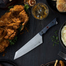 The Messermeister Iron & Resin Overland Chef 8″ Camp Knife by Messermeister rests on a dark tabletop near fried chicken, a glass of whiskey, mashed potatoes, biscuits, and herbs, creating a rustic meal scene.