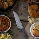 The Messermeister Iron & Resin Overland Chef 8″ Camp Knife by Messermeister, featuring a precision-forged blade, rests on a wooden table surrounded by baked beans, potato salad, fried chicken, ribs, potato chips, a sandwich, and breaded food in a skillet—making it the ultimate companion for both outdoor cooking and backyard feasts.