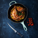 A cast iron pan filled with large shrimp atop a bed of diced vegetables, garnished with parsley. Nearby on a dark blue surface rests the Messermeister Iron & Resin Overland Chef 7″ Mid-Flex Fillet Knife by Messermeister, ideal for fish preparation thanks to its high-carbon stainless steel blade, along with a vine of cherry tomatoes.