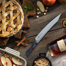The Messermeister Iron & Resin Overland Chef 9″ Carving Knife rests on a rustic wooden table, surrounded by apple pie, brown sugar, sliced apples, cinnamon sticks, spices, and a bottle of liquor—setting a cozy autumn baking scene.