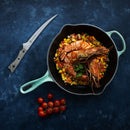 A cast iron pan with large cooked shrimp atop a bed of diced vegetables rests on a blue surface. Nearby sit a bunch of cherry tomatoes and a knife from the Messermeister Overland Chef 6-Piece Leather Knife Roll Set—essentials for anyone who values quality outdoor cooking knives from Messermeister.