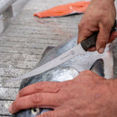 A person fillets a fish on a cutting board using a long, thin knife. Nearby, a Messermeister Overland Chef 6-Piece Leather Knife Roll Set is open, showcasing an array of Messermeister outdoor cooking knives. In the background, there is a piece of salmon fillet.