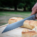 A hand uses a serrated knife from the Messermeister Overland Chef 6-Piece Leather Knife Roll Set to slice a baguette on a wooden cutting board outdoors, with grass in the background.