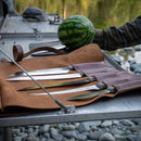 The Messermeister Overland Chef 6-Piece Leather Knife Roll Set is displayed open on a metal table outdoors, showcasing several large outdoor cooking knives. Nearby, a person holds a whole watermelon, with a river and rocks visible in the background.