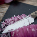 A close-up of a Messermeister Overland Chef Utility Knife - 4.5", expertly crafted from premium Nitro cobalt steel, on a cutting board with chopped red onion pieces and part of a whole red onion in the foreground.