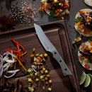 A knife from the Messermeister Overland Chef 6-Piece Leather Knife Roll Set rests on a wooden cutting board beside sliced onions and peppers, surrounded by tacos filled with grilled meat, vegetables, and fresh herbs.