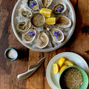 A plate of fresh oysters on ice with edible flowers, lemon wedges, and mignonette sauce sits beside extra sauce, salt, and a Messermeister Oyster Knife - 6.5" by Messermeister on the wooden table.