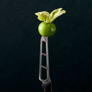A fresh green tomatillo with its husk partially open is balanced on the tip of a Messermeister Petite Messer Black Cheese & Tomato Knife - 6" against a dark, textured background.