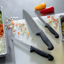 The Messermeister Pro Series Wide Chef's Knife - 8" by Messermeister, with a black handle, sits on a white cutting board beside diced carrots, celery, onion, and yellow and red peppers—perfect for precise prep in any kitchen.