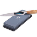 A Messermeister kitchen knife with a wooden handle rests on the Messermeister Restoration Stone - 400 & 1000 Grit, a rectangular gray sharpening stone offering dual grit surfaces for precise blade maintenance.