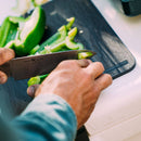 Close-up of hands chopping green bell peppers with a knife on the Messermeister Adventure Chef Folding Faux Wood Cutting Board. The chef, wearing a blue long-sleeve shirt, preps ingredients for the camp kitchen.