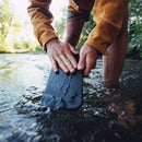 A person kneels in a stream, panning for gold with a black pan. Nearby, the Messermeister Adventure Chef Folding Faux Wood Cutting Board adds convenience to their camp kitchen amid greenery and sunlight.