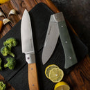 The Messermeister Adventure Chef Linen Folding Chef's Knife - 6" rests on a black cutting board, surrounded by lemon slices, broccoli florets, garlic cloves, and a yellow bell pepper arranged on a wooden surface.