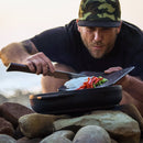 A man in a camouflage cap uses the Messermeister Adventure Chef Maple Basecamp Set - 3 Piece to slice vegetables on a cutting board and transfer them into a black pot outdoors, with rocks visible in the foreground.