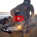 A man in a Cook Outside hat uses the Messermeister Adventure Chef Maple Folding Camp Utensil to pour oil onto food cooking over a beach campfire at sunset. His dog rests nearby, and a play button overlay is displayed.